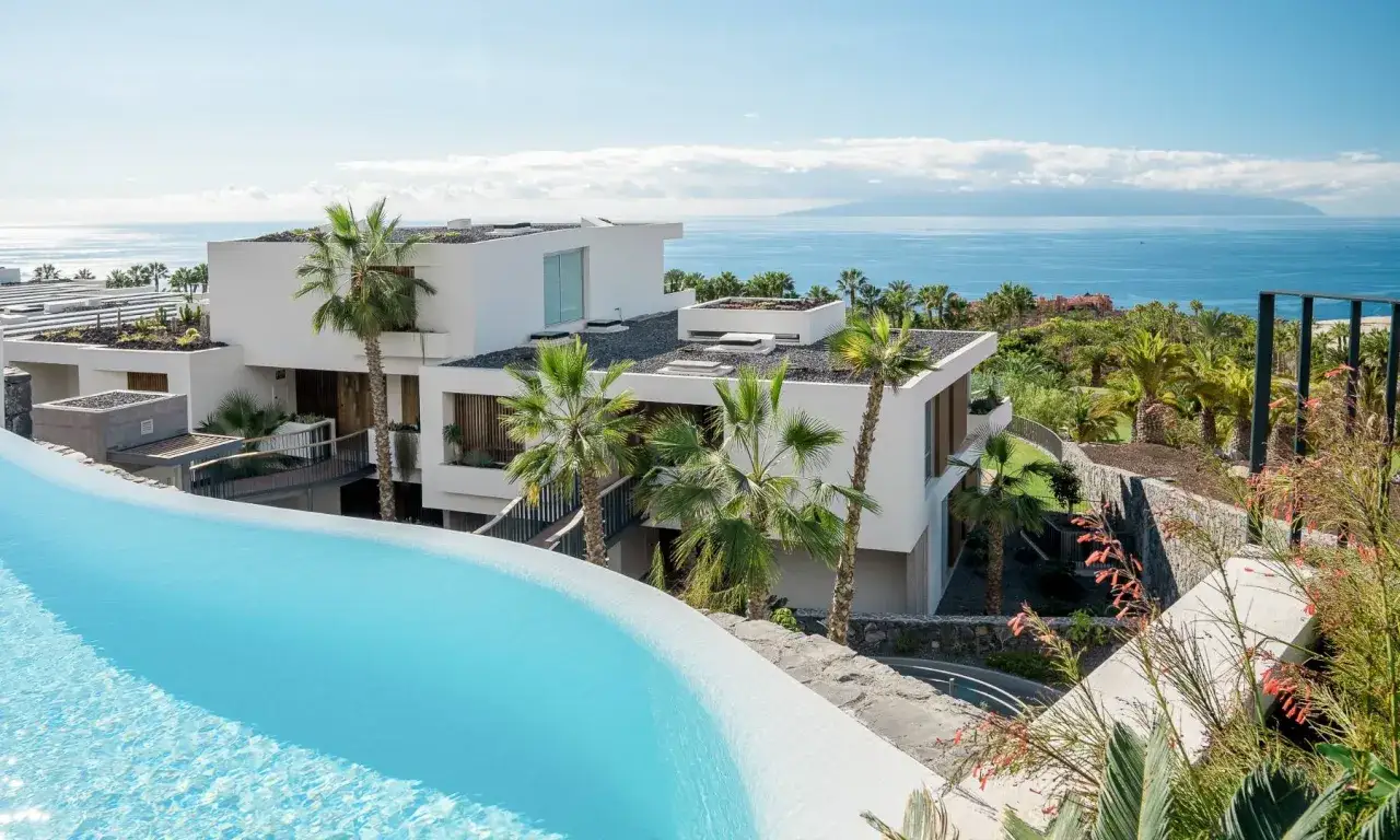 Piscina infinita con vistas al mar y arquitectura moderna en Gran Meliá Palacio de Isora. Palmeras y vegetación exuberante.