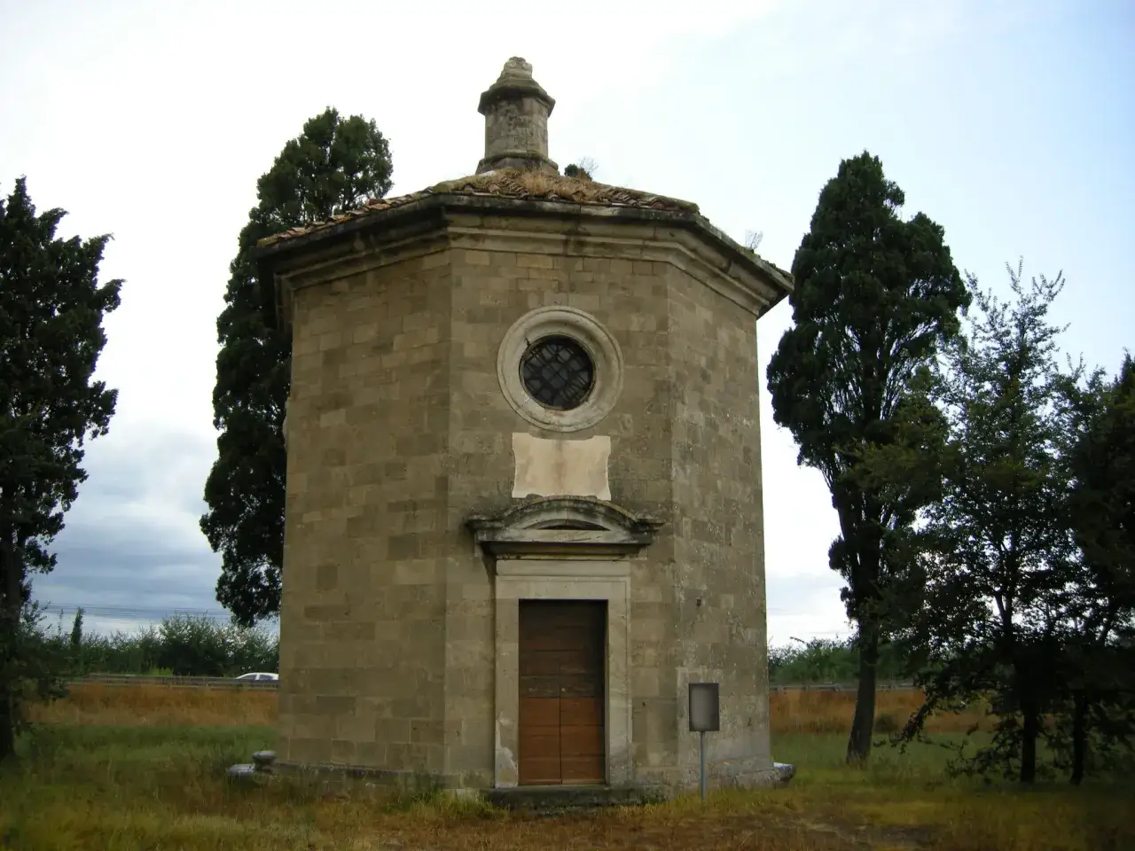 Piccola cappella ottagonale in pietra, circondata da alberi, su un viale dei cipressi in Toscana.