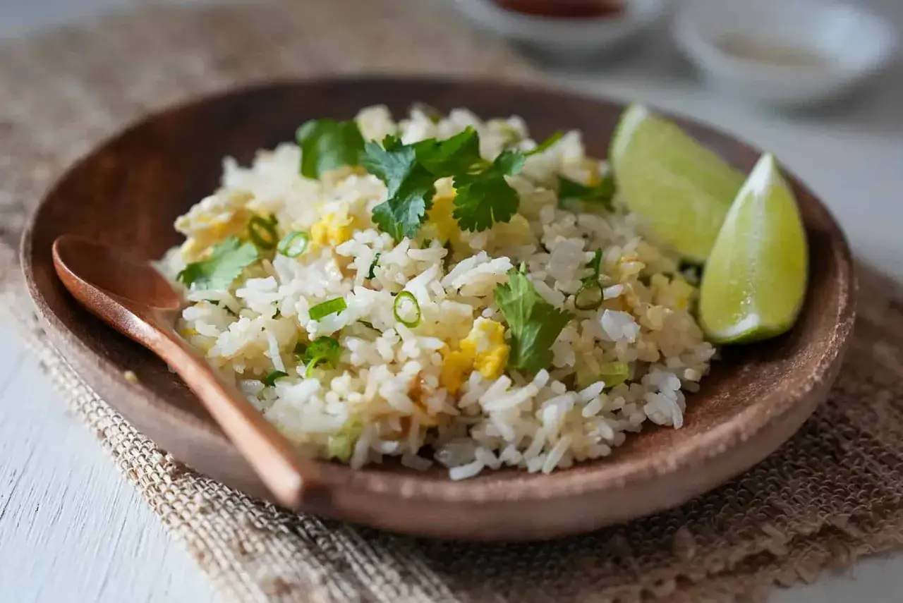 En skål med khao pad, en thailändsk stekt risrätt, garnerad med koriander och limeklyftor. En träsked ligger bredvid.