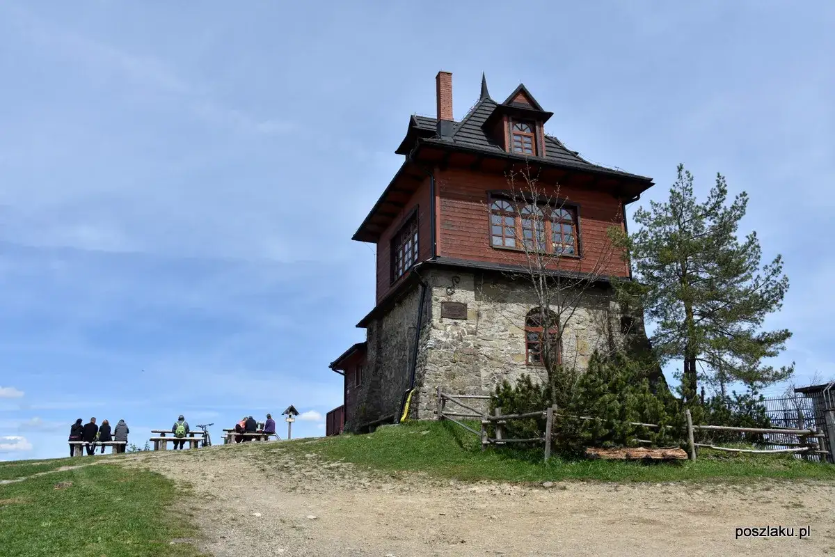 Drewniane schronisko na Luboniu Wielkim, z kamiennym fundamentem. Ludzie odpoczywają na ławkach na tle błękitnego nieba.
