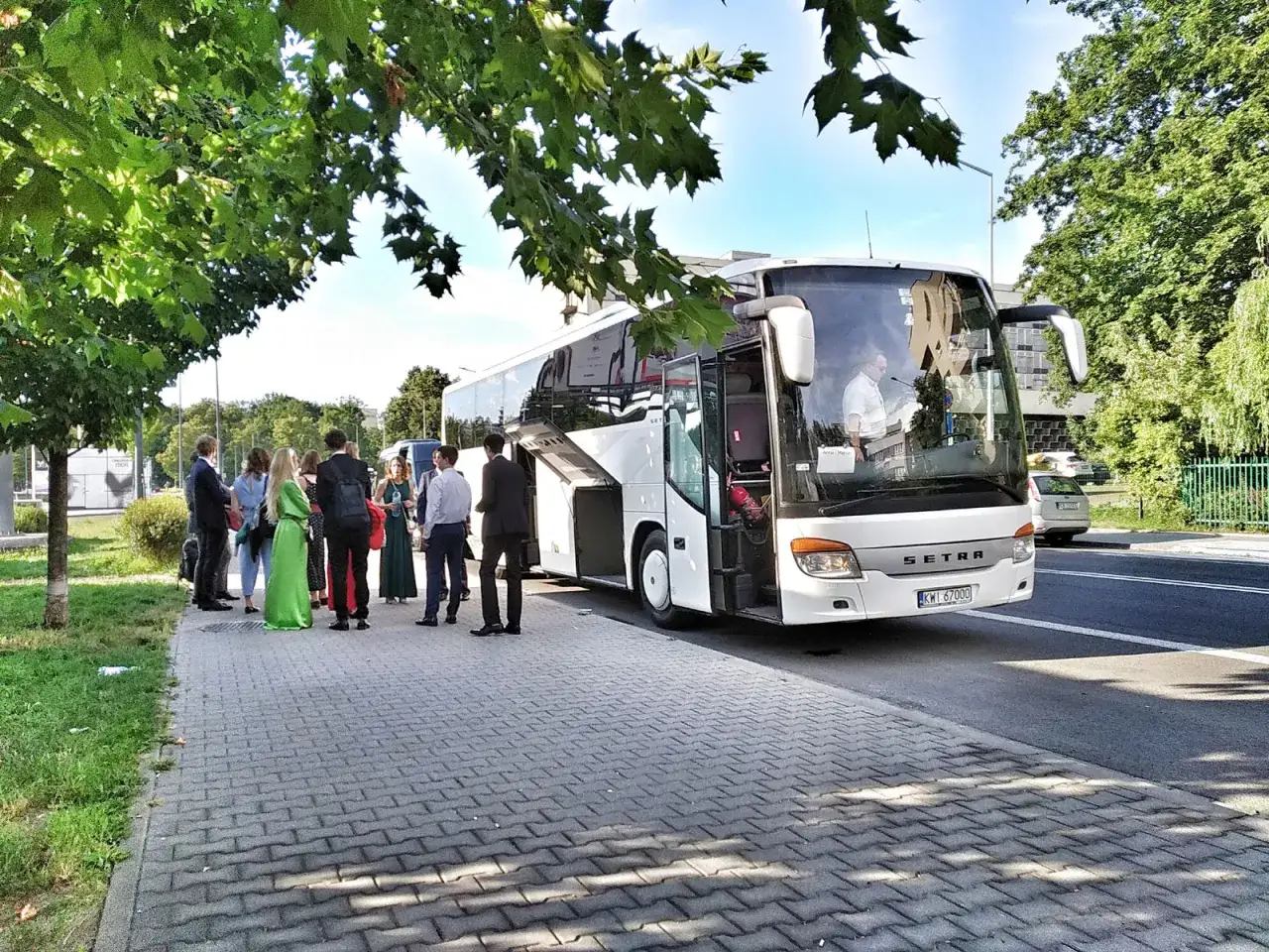 Elegancko ubrani goście weselni czekają na transport. Biały autokar Setra z otwartymi drzwiami czeka, by zabrać ich na uroczystość.