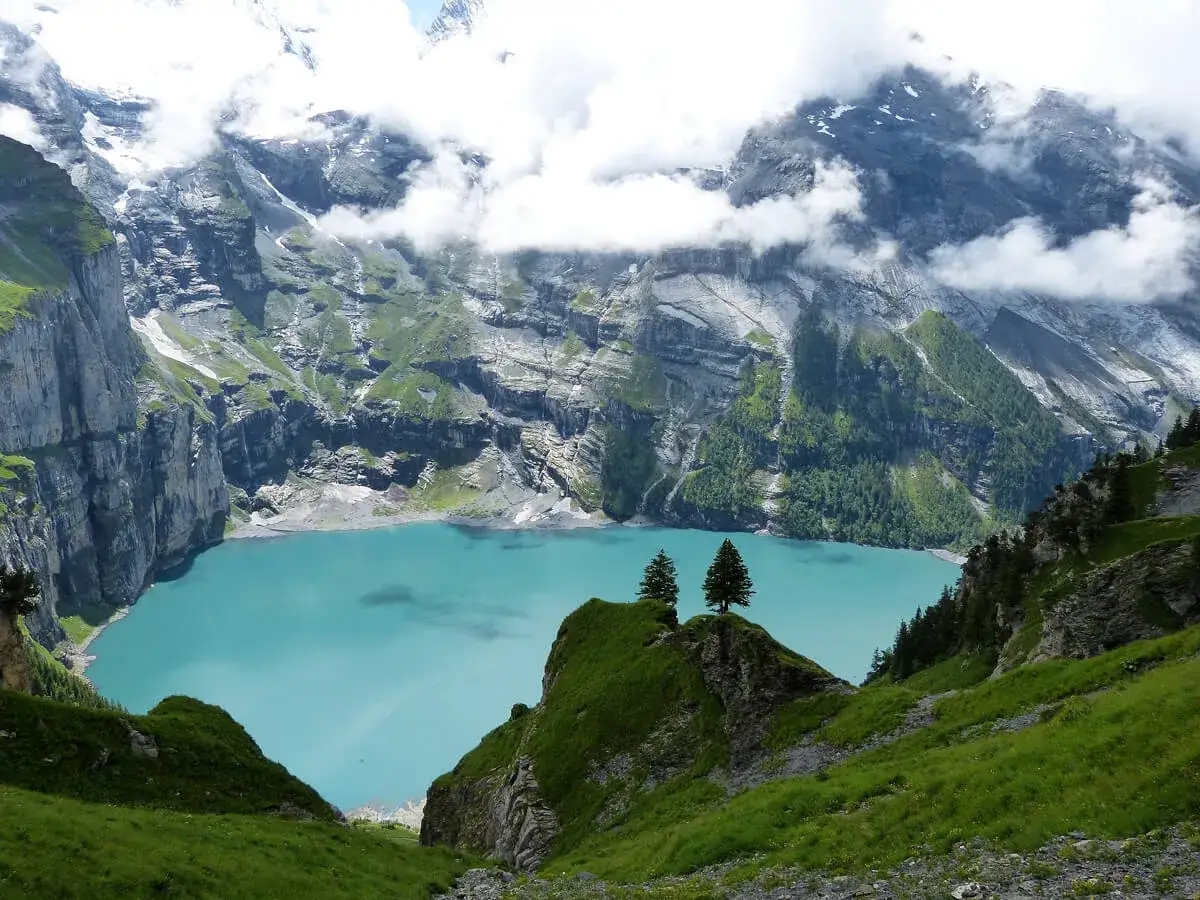 T&uuml;rkisfarbener See Oeschinensee Schweiz, umgeben von steilen Felsw&auml;nden und gr&uuml;nen H&auml;ngen, mit Wolken, die die Berggipfel umh&uuml;llen.