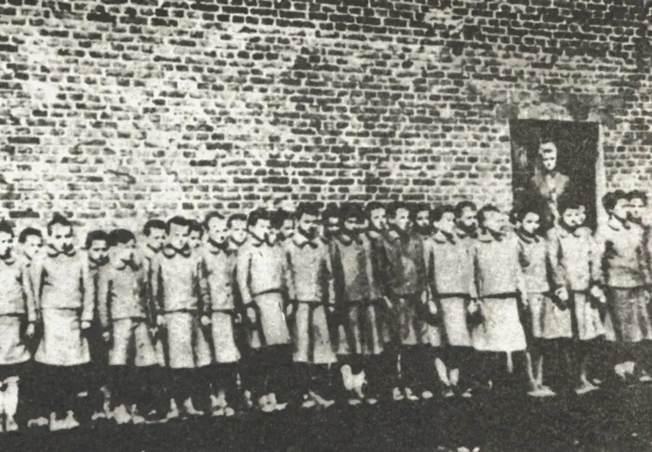 Barn i en rad framf&ouml;r en tegelv&auml;gg, en m&ouml;rk skugga av en vuxen syns i en d&ouml;rr&ouml;ppning. En sorglig p&aring;minnelse om hitler barn.