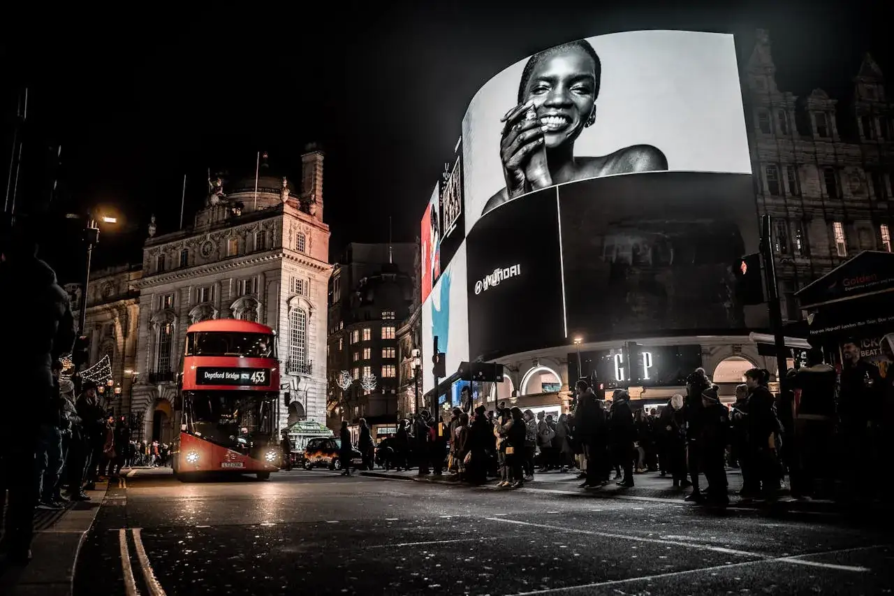 Nocne światła Piccadilly Circus, gdzie specjalista ds reklamy podziwia ogromne billboardy. Czerwony autobus i tłum ludzi.
