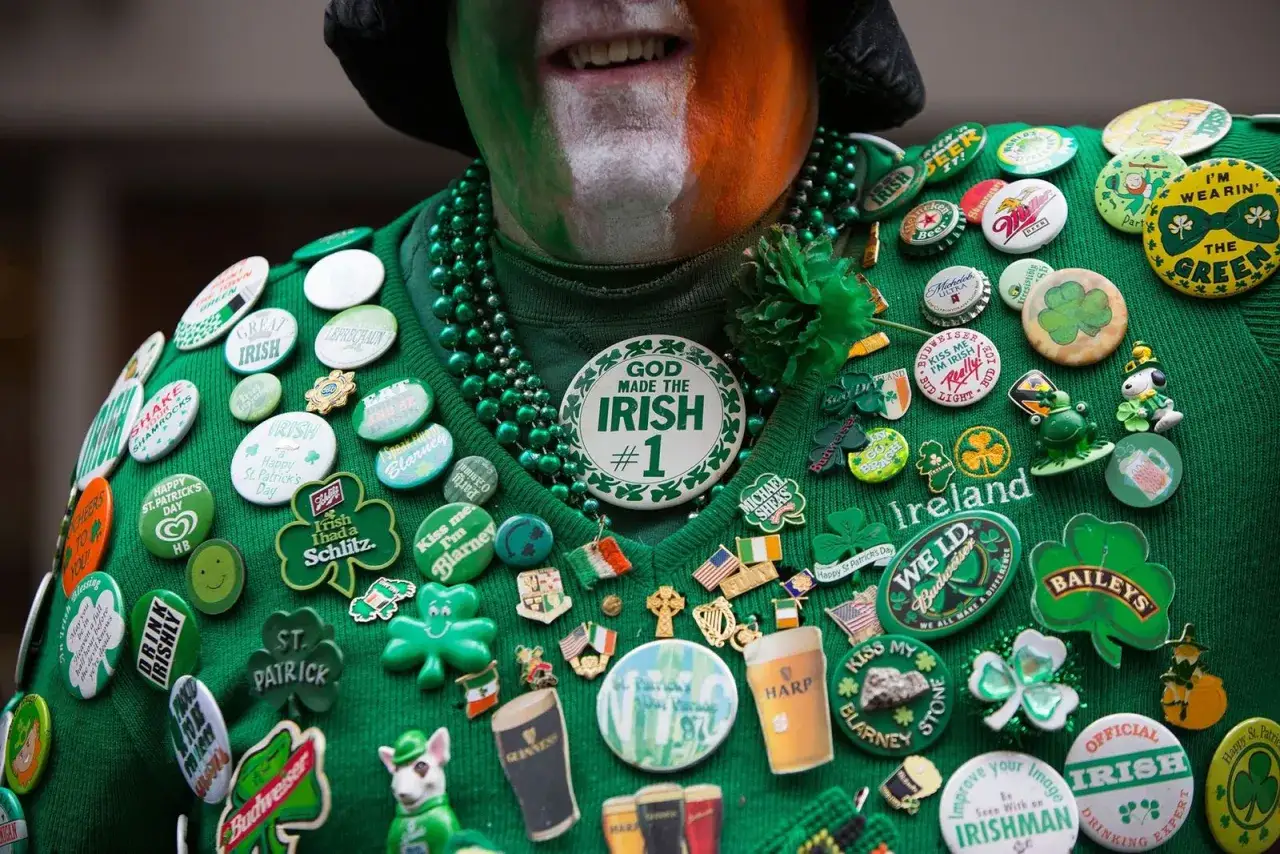 Un hombre celebra el Día de San Patricio con un suéter verde cubierto de insignias, collares y pintura facial tricolor.