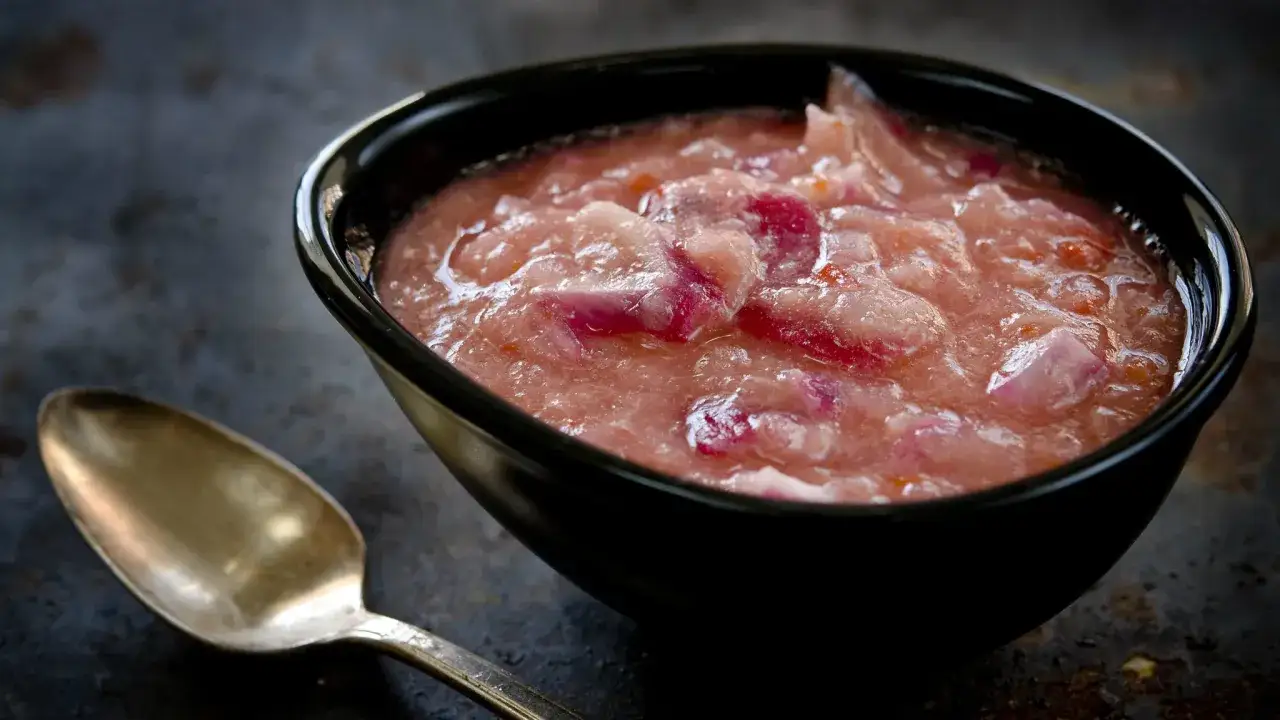 En sk&aring;l med rabarber chutney, med bitar av rabarber och r&ouml;dl&ouml;k, bredvid en gammal sked.