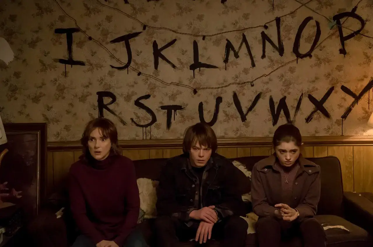 Joyce Byers i jej dzieci, Jonathan i Nancy, siedzą na kanapie, patrząc na ściany z namalowanymi literami, jak w Stranger Things 1.