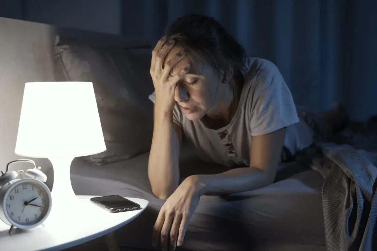 Kobieta leży w łóżku, trzymając się za czoło. Mimo zmęczenia nie może zasnąć. Obok stoi lampka, budzik i telefon.