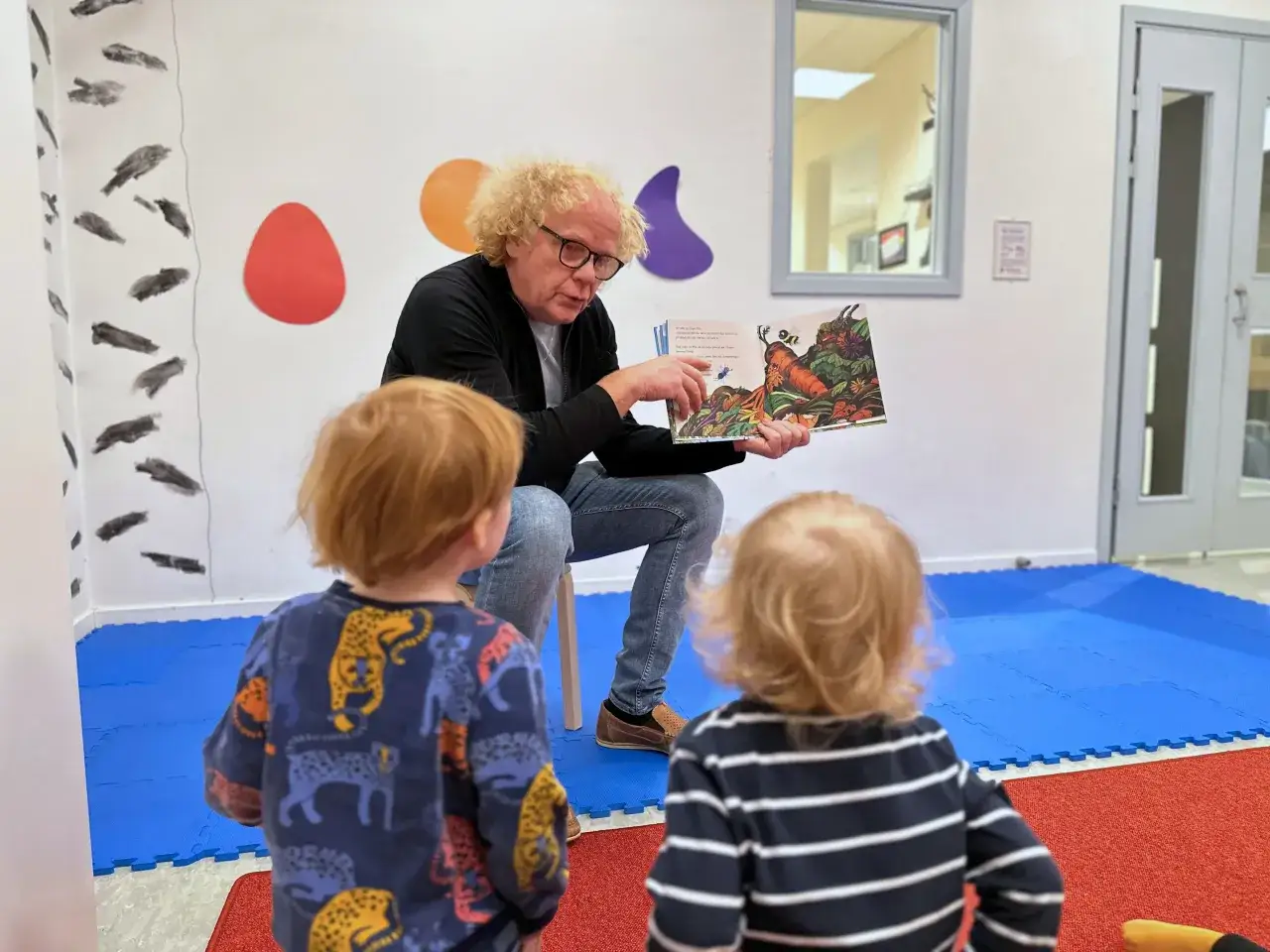 En man med lockigt hår läser en bok för två små barn. Bilden visar en humlan djojj på en morot i boken.