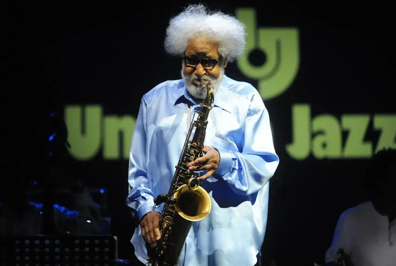 Un sassofonista jazz con una folta chioma bianca suona il suo strumento su un palco illuminato.
