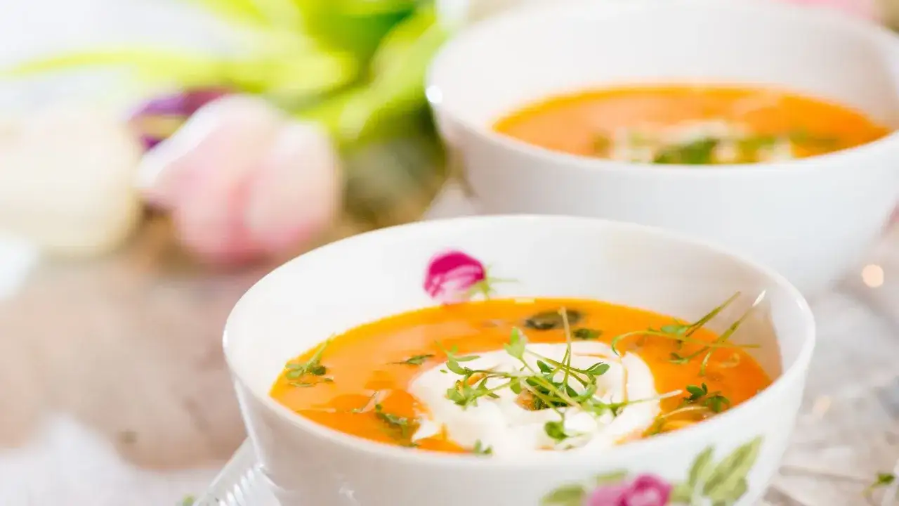 Zwei Schalen cremiger Karottensuppe, garniert mit frischen Kr&auml;utern und einem Klecks saurer Sahne, vor fr&uuml;hlingshaften Blumen.