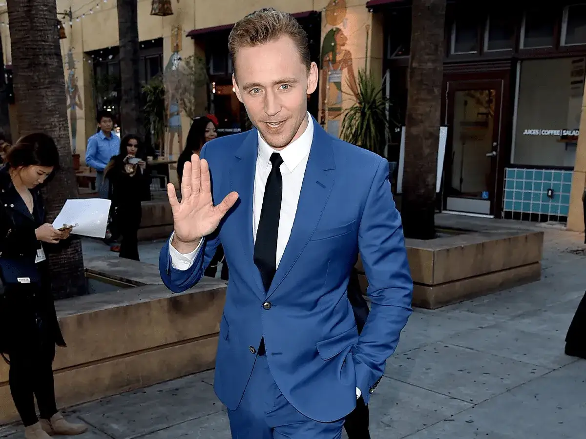 Tom Hiddleston waves, sporting sharp navy blue suit combinations with a white shirt and black tie.