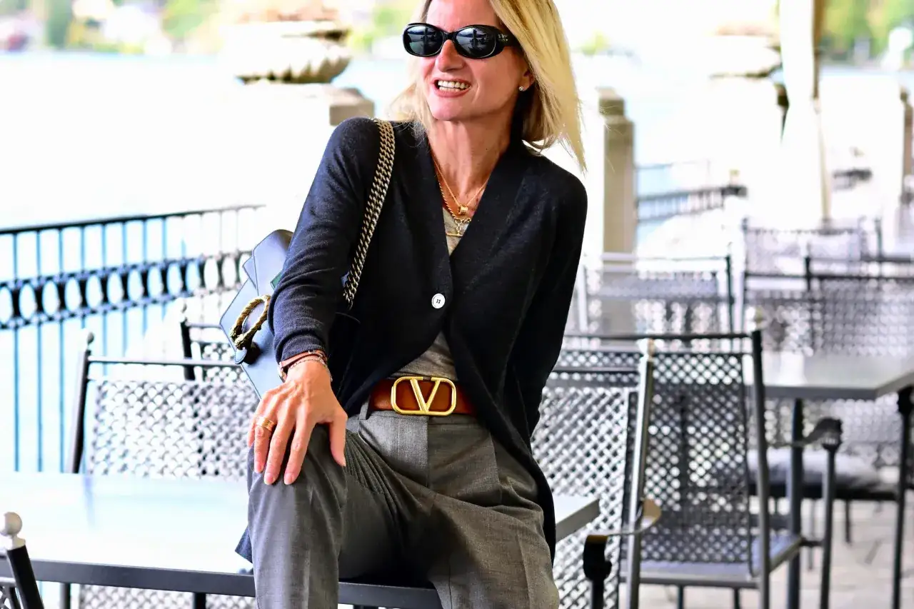 Donna con pantalone grigio elegante, abbinato a cardigan scuro e cintura con logo dorato. Un look chic per ogni occasione.