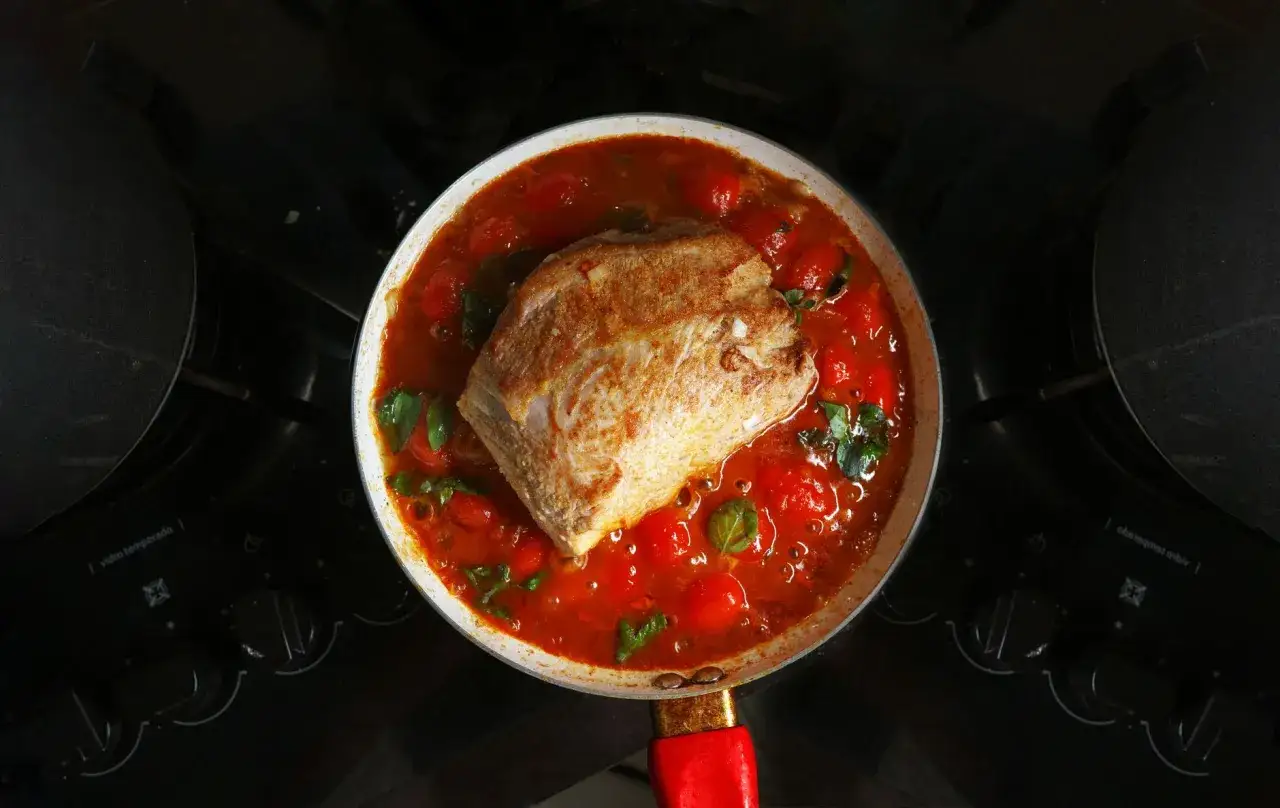 Un morceau de sauté de porc mijote dans une sauce tomate avec des tomates cerises et du basilic, dans une poêle sur une cuisinière.
