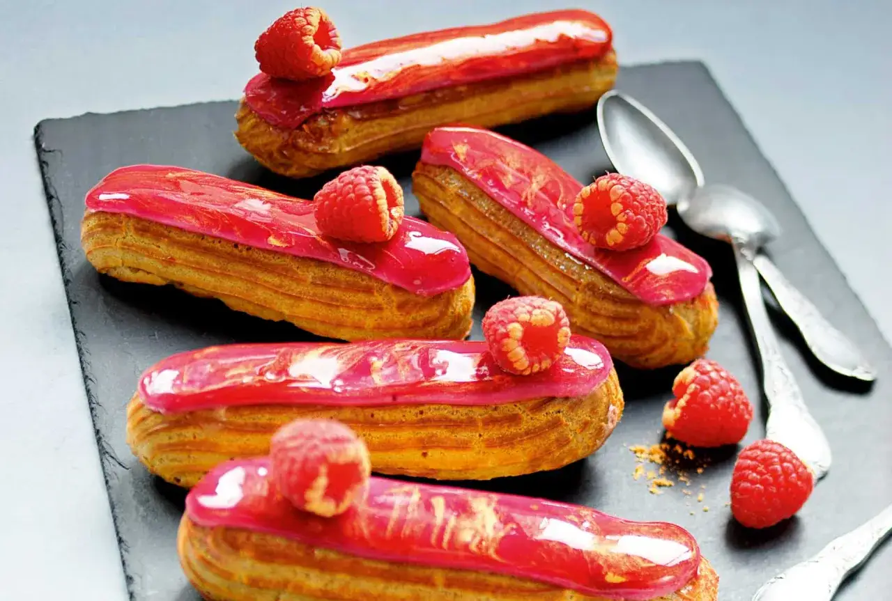 Cinq &eacute;clairs &agrave; la framboise, glac&eacute;s de rose vif et orn&eacute;s de fruits rouges frais, sur une ardoise.