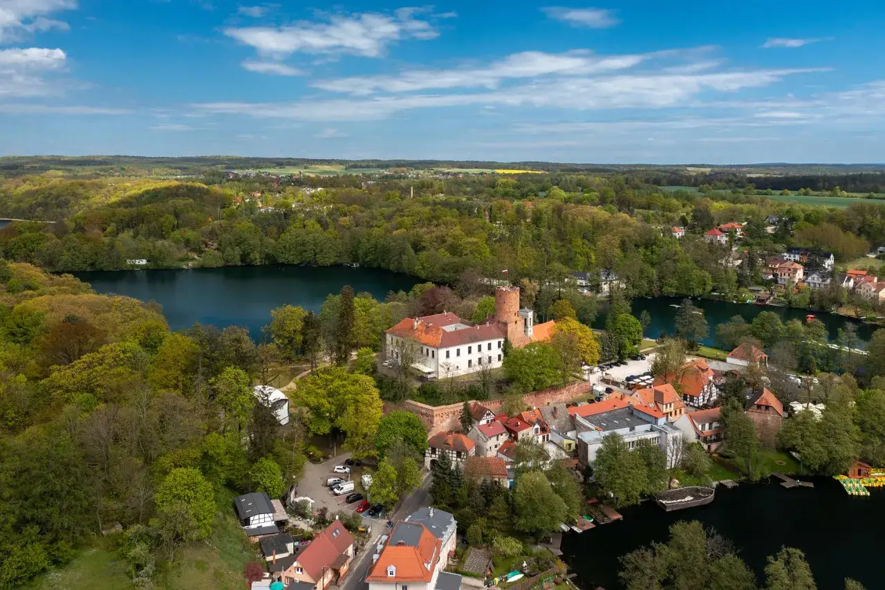 Malowniczy widok na zamek i zabudowania nad jeziorem Łag&oacute;w, otoczone bujną zielenią drzew.