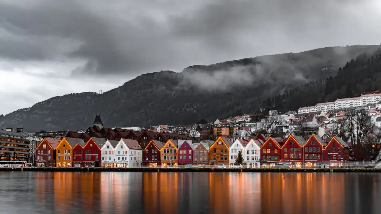 Kolorowe domy w Bryggen, Bergen, Norwegia, odbijające się w wodzie. W tle g&oacute;ry i chmury. Uważaj na mandaty w Norwegii, jeśli parkujesz nieprawidłowo.