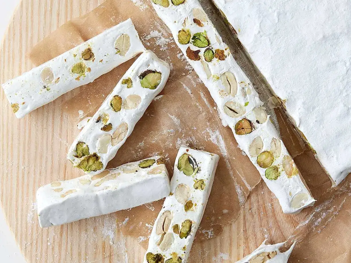 Flera bitar fransk nougat med pistagen&ouml;tter och mandlar ligger utspridda p&aring; ett tr&auml;underlag.