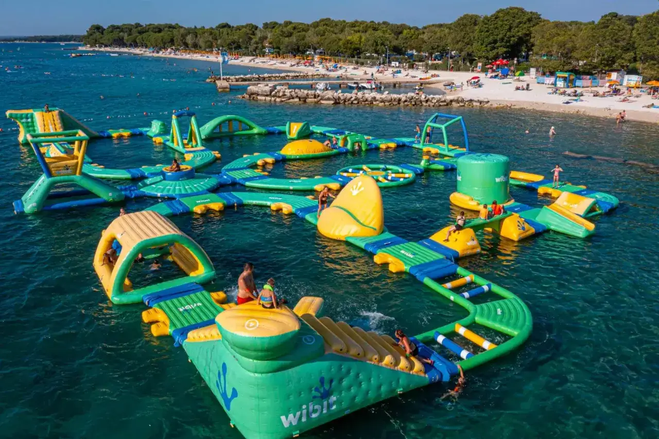 Wesołe miasteczko na wodzie w bi village camping. Dzieci i dorośli bawią się na dmuchanych zjeżdżalniach i torach przeszkód w turkusowej wodzie.