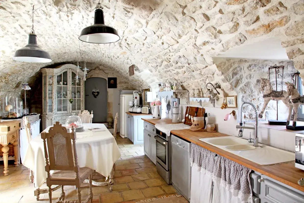Cuisine rustique avec des vieux meubles patinés, murs en pierre, vaisselle ancienne et une table dressée.