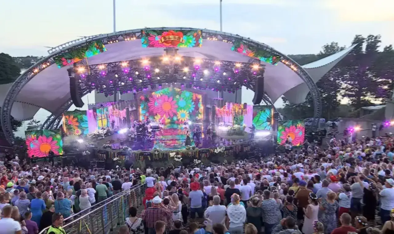 Koncert zespołów disco polo na scenie z kolorowymi kwiatami. Tłumy bawią się pod gołym niebem.