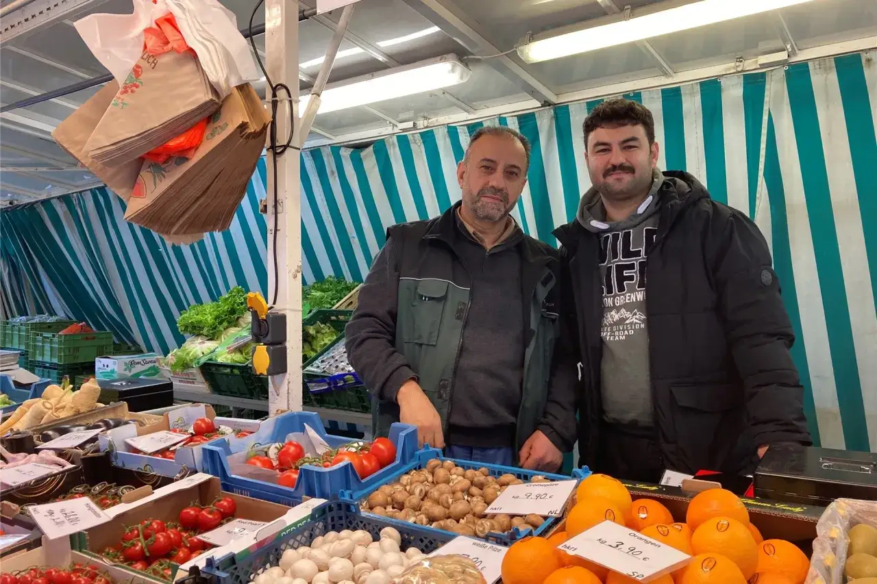 Zwei Männer stehen an einem Obst- und Gemüsestand auf dem markt kreis recklinghausen. Frische Tomaten, Pilze und Orangen sind zu sehen.