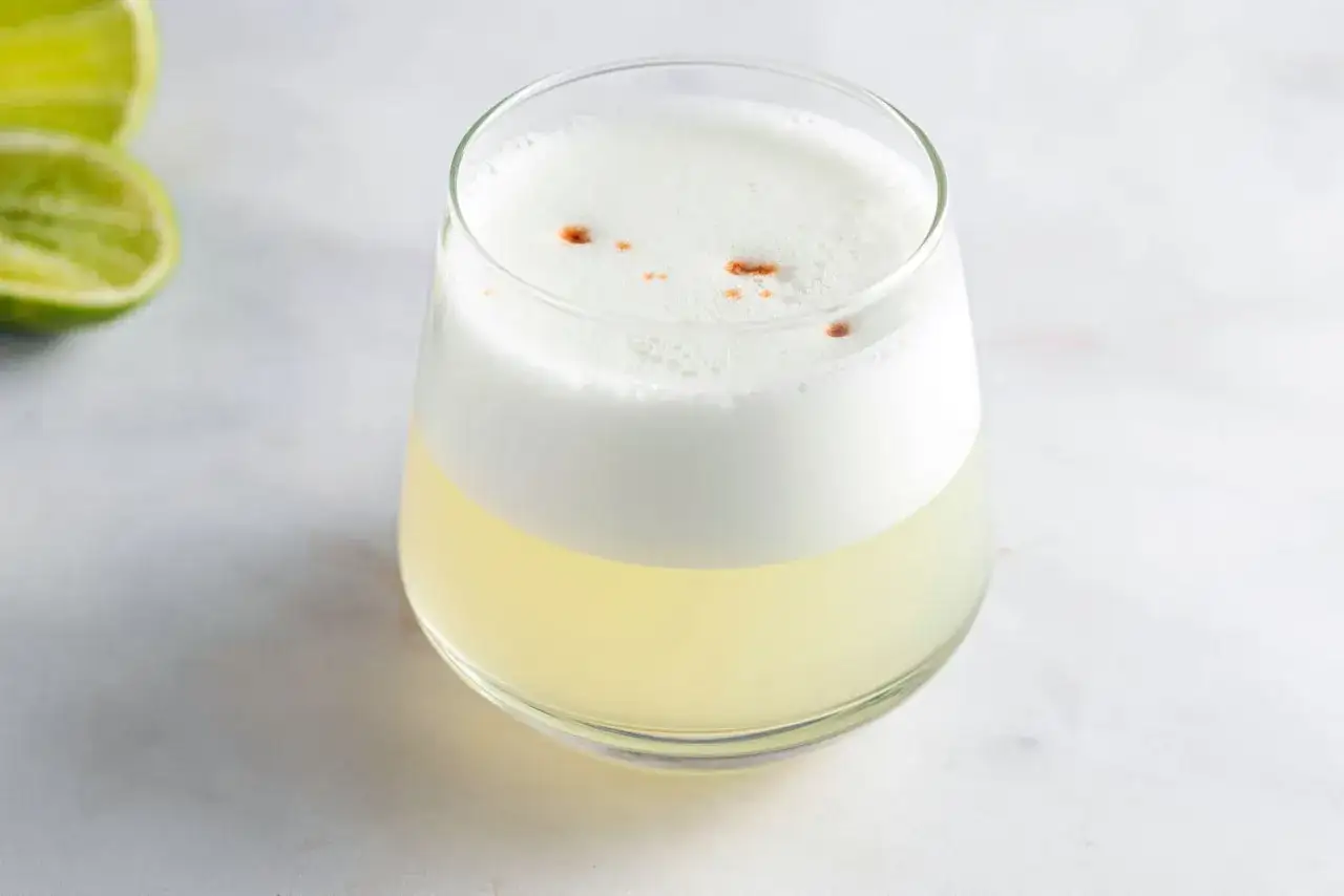 En glas pisco sour med skummig topp och en nypa kanel. Två limeklyftor ligger till vänster.