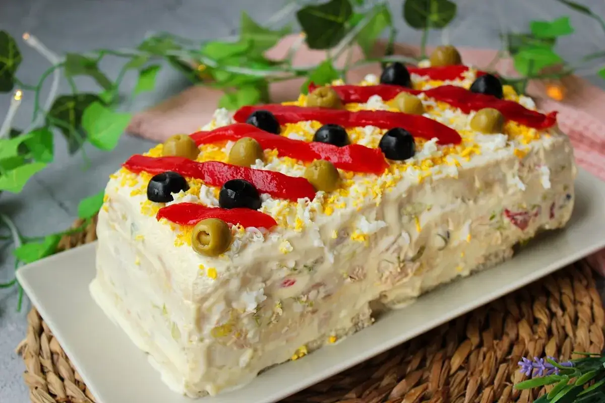 Pastel de at&uacute;n con pan de molde, decorado con pimientos rojos, aceitunas negras y verdes, y huevo rallado. &iexcl;Una delicia para compartir!