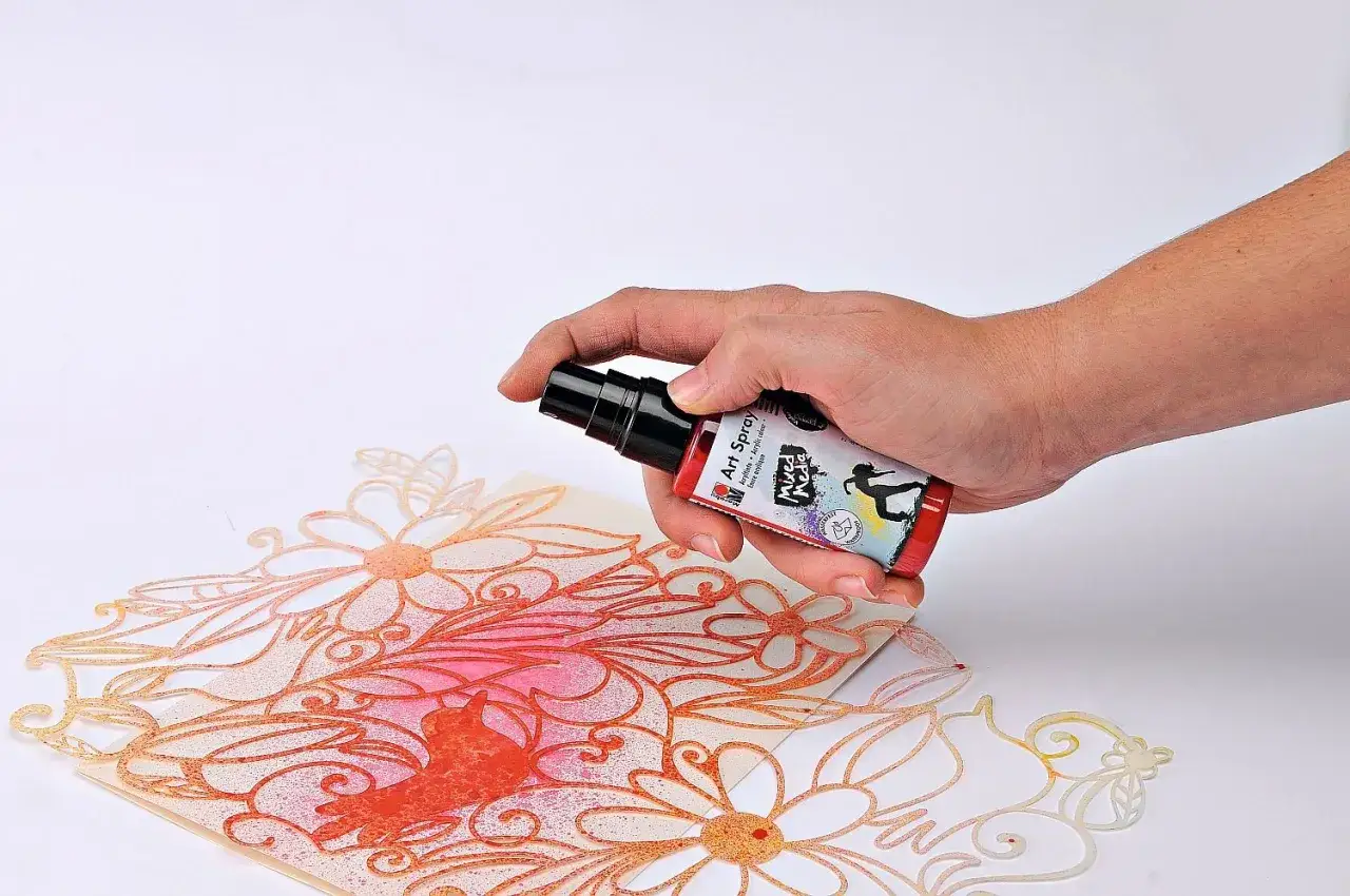 Main tenant une bombe de peinture rouge, pulv&eacute;risant sur un motif floral d&eacute;coup&eacute; sur du carton.