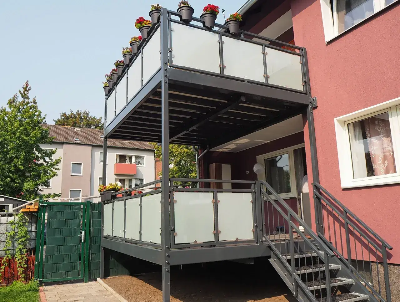 Nowoczesny, dwupoziomowy balkon dostawny do domu jednorodzinnego, z metalową konstrukcją, szklanymi balustradami i kwitnącymi kwiatami w donicach.