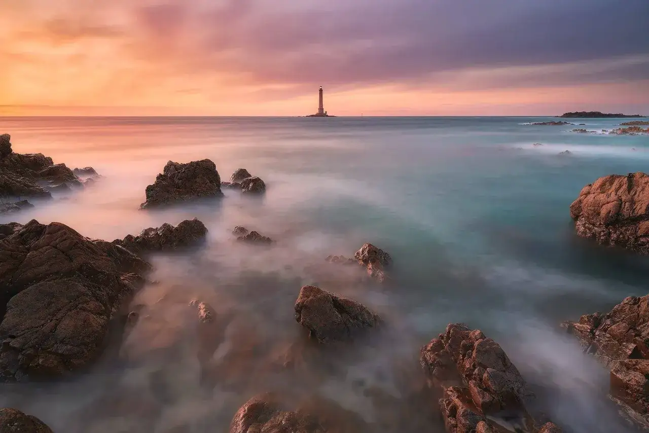 Un phare se dresse au loin sur une &icirc;le rocheuse, baign&eacute; par les couleurs douces d'un coucher de soleil. L'eau, rendue soyeuse par un filtre ND, caresse les rochers au premier plan.