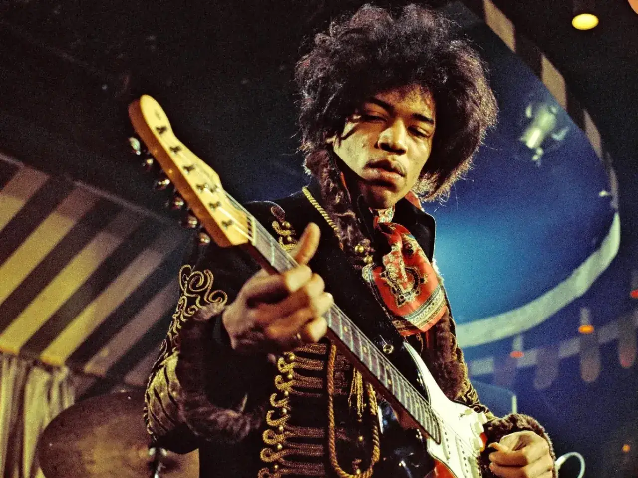 Jimi Hendrix, icône du groupe, joue de sa guitare électrique avec passion sur scène, vêtu d'une tenue ornée.