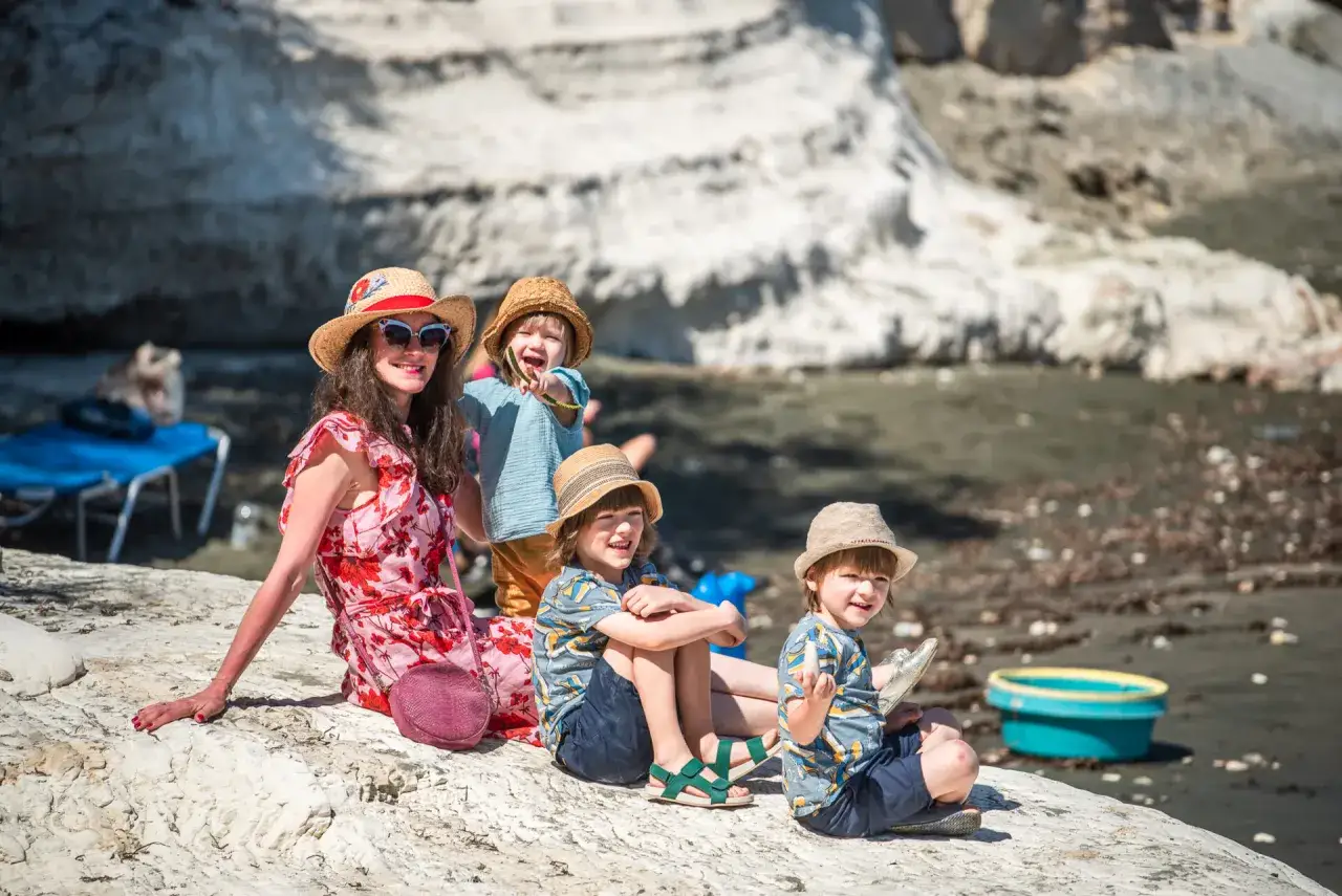 Rodzina cieszy się wakacjami z dzieckiem na plaży. Mama i tr&oacute;jka dzieci w słomkowych kapeluszach siedzą na kamieniach.