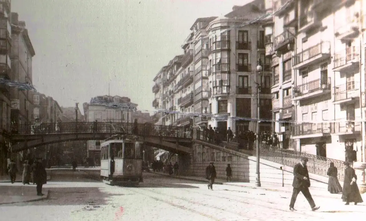 Un tranv&iacute;a cruza la calle bajo el puente de Santander, rodeado de edificios antiguos y gente paseando.