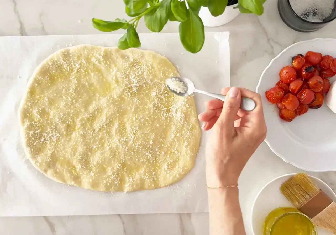 Eine Hand streut Parmesan auf einen rohen Teig für italienische Sauerteig Pizza. Daneben liegen geröstete Kirschtomaten.