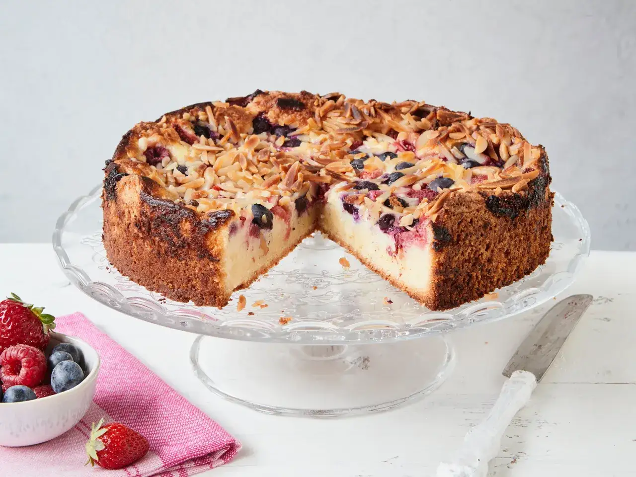 Ein saftiger **Beerenkuchen vom Blech** mit Mandelsplittern, serviert auf einem Tortenständer. Daneben eine Schale mit frischen Beeren.