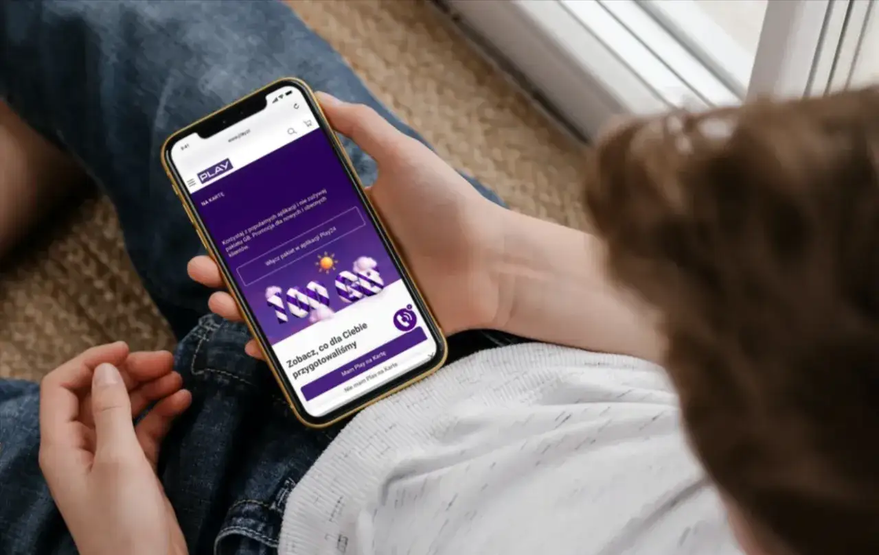 Dłoń trzyma smartfon z aplikacją Play, gdzie widnieje informacja o promocji i pakietach. To ułatwia automatyczny wybór sieci Play.