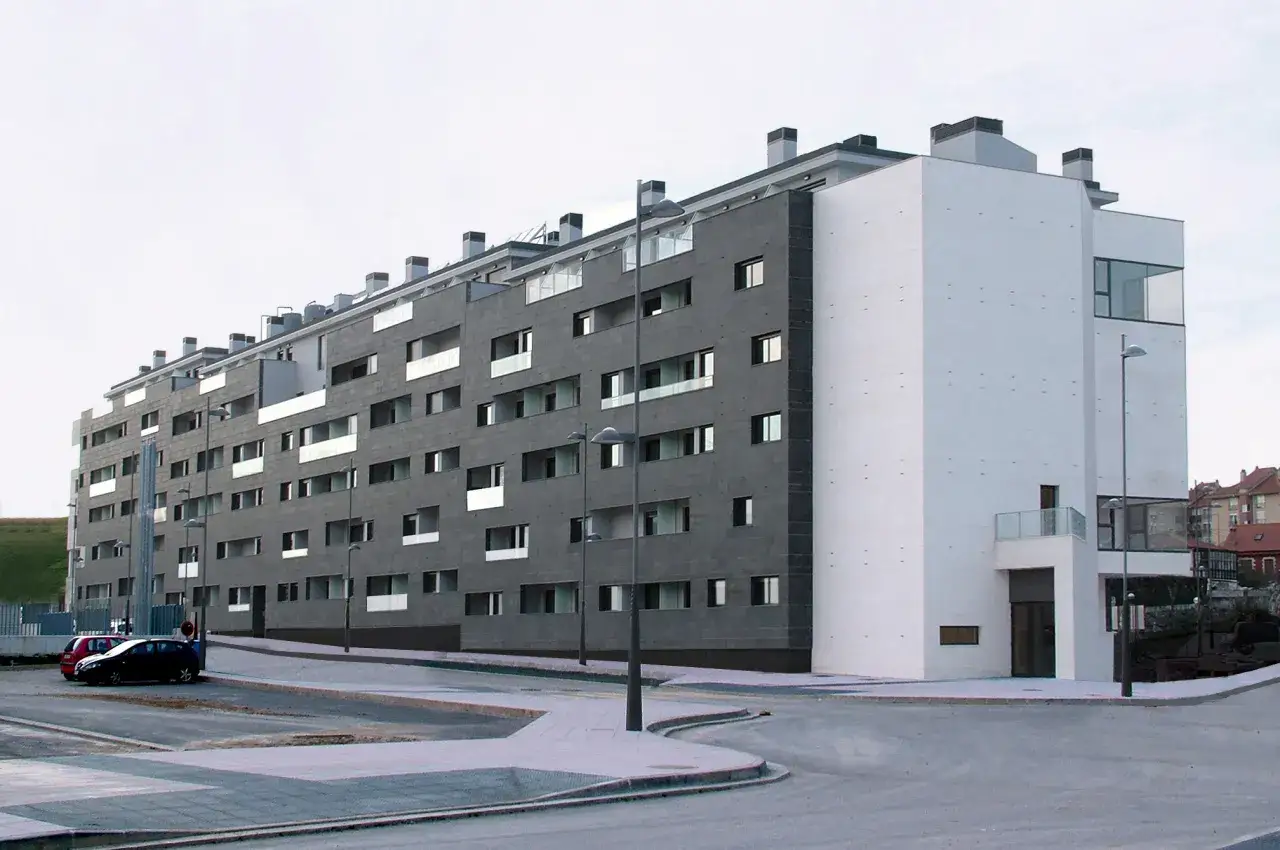 Edificio moderno con fachada de piedra oscura y blanca. Hay coches aparcados y farolas. Si no cumples las normas, te pueden echar de una vivienda de protección oficial.