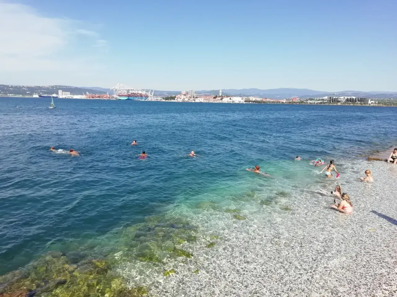 Ludzie kąpią się w krystalicznie czystej wodzie przy plaży w Koprze, Słowenia. W tle widać port i miasto.