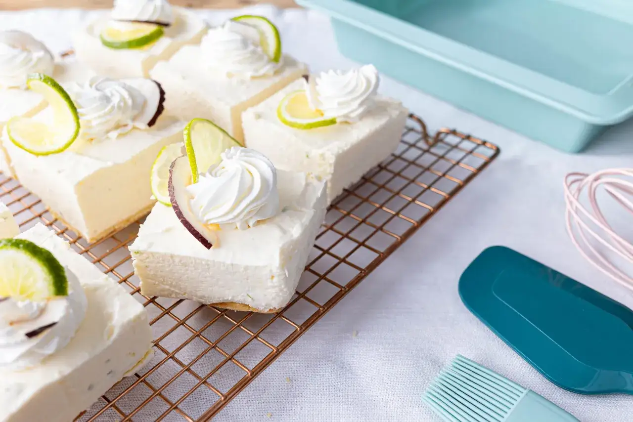 Fette di cheesecake al cocco, guarnite con panna montata e fettine di lime, riposano su una griglia.