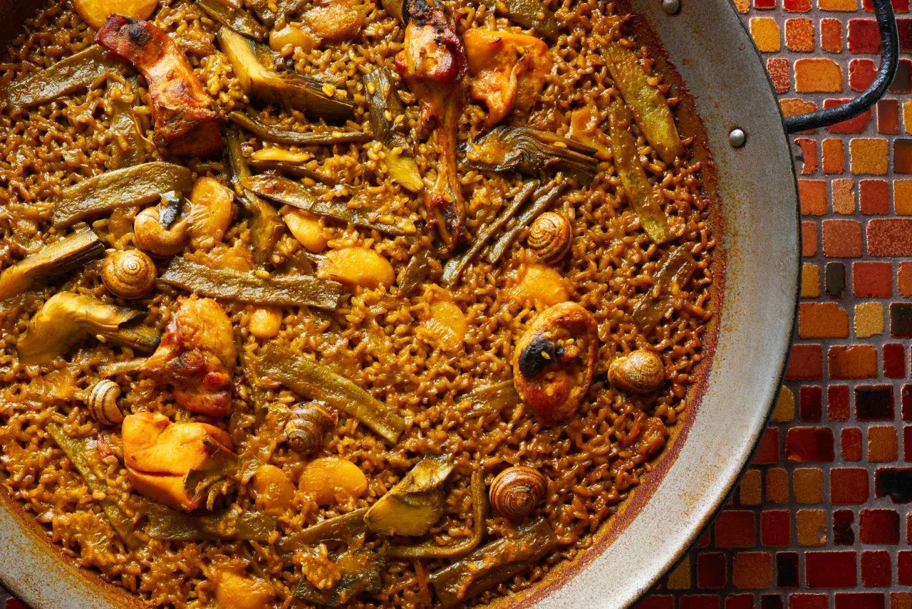 Paella de pollo con trozos de pollo dorado, jud&iacute;as verdes, alcachofas y caracoles, servida en una paellera sobre un mosaico de colores.