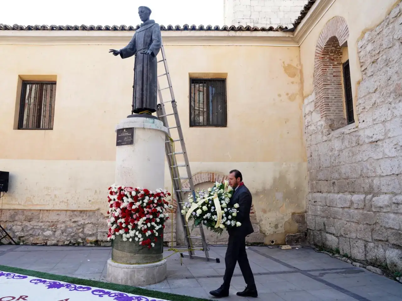Hombre con corona de flores se acerca a la estatua de San Pedro Regalado, adornada con flores rojas y blancas.