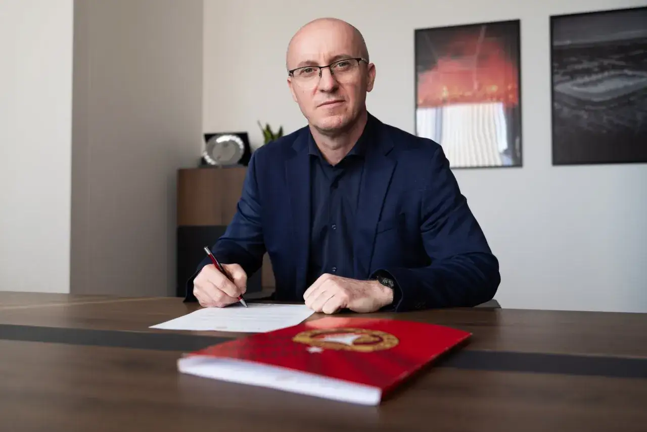 Nowy trener Widzewa podpisuje kontrakt. Mężczyzna w garniturze siedzi przy biurku, pisząc piórem.