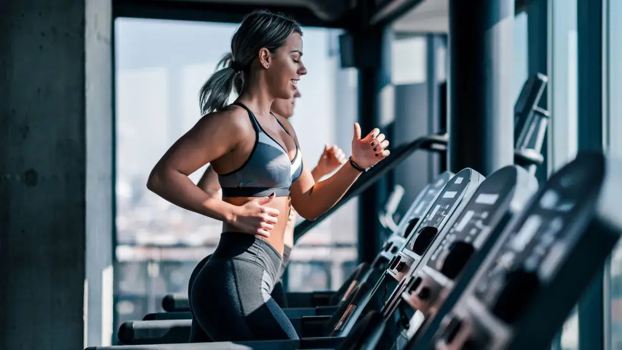Uśmiechnięta kobieta podczas treningu cardio na siłowni, biegnie na bieżni obok innej osoby.