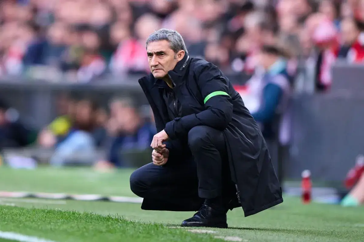 Ernesto Valverde, w czarnej kurtce z zielonym pasem, kuca na murawie, skupiony na prowadzonej drużynie.