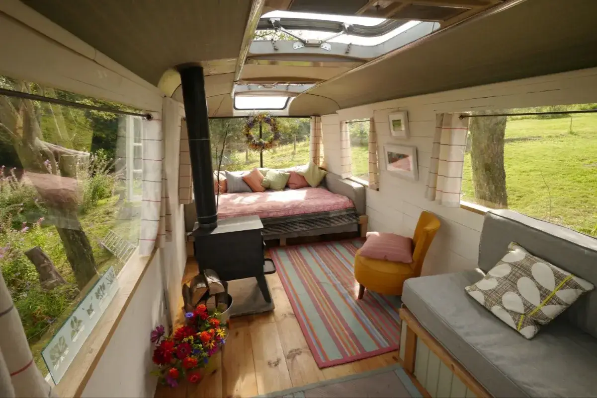 Interior acogedor para decorar caravana por dentro: cama, estufa de leña, sillón amarillo y vistas al campo.