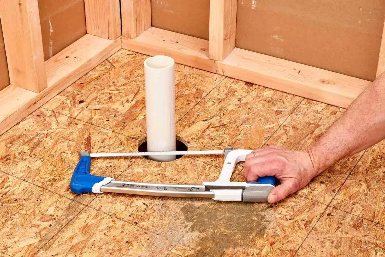 A hand uses a hacksaw to cut a hole in the subfloor for a shower pan installation, with a drain pipe visible.