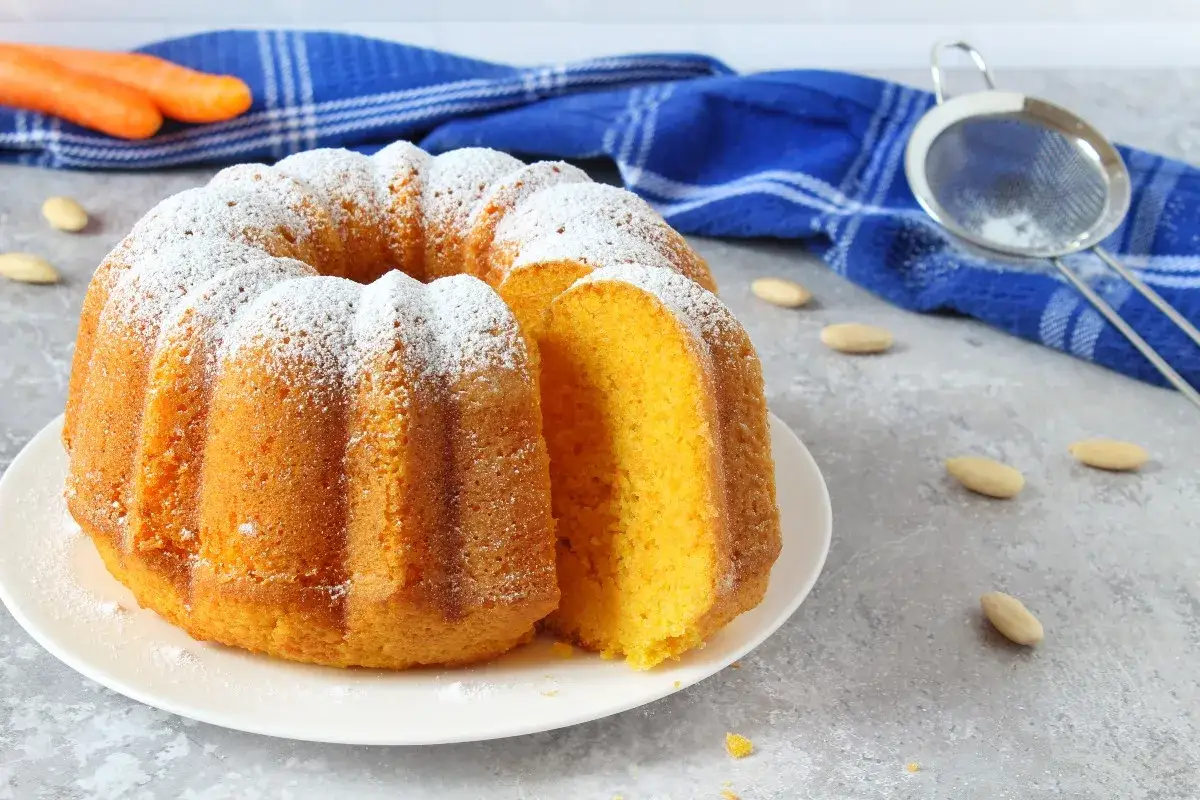 Torta carote e mandorle soffice e profumata, cosparsa di zucchero a velo. Sullo sfondo, carote fresche e mandorle.