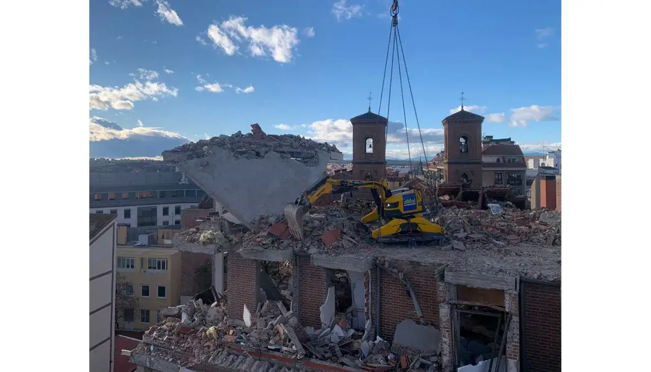 Excavadora amarilla en medio de demoliciones de edificios, con grúa y torres de iglesia al fondo.
