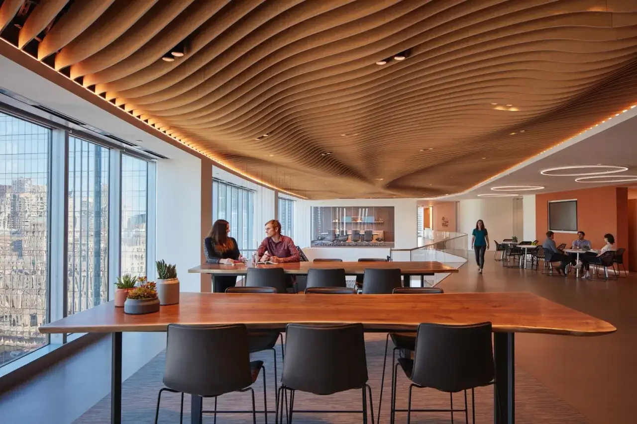 Un **open space** moderne avec un plafond ondul&eacute; en bois. Deux personnes discutent &agrave; une table pr&egrave;s d'une grande fen&ecirc;tre.